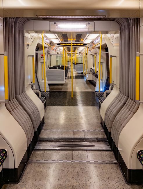 The interior of a modern train carriage showing clean, smooth surfaces with beige and grey plastic seats arranged along both sides facing inward, separated by a central aisle with a speckled grey floo