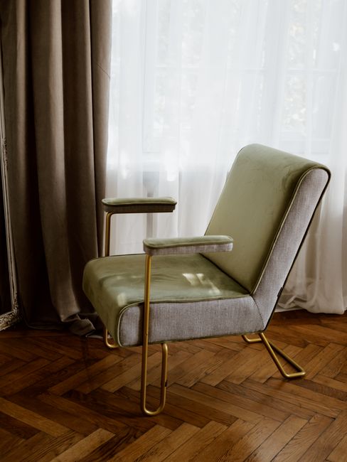 A beige and light green upholstered armchair with gold-colored metal armrests and legs, positioned on a polished wooden herringbone-patterned floor next to a window dressed with sheer white curtains and heavier brown drapes. Natural light filters through the curtains, illuminating the clean and well-maintained domestic room. The setting suggests a focus on home surface cleaning and furniture maintenance, with the armchair appearing freshly cleaned and free of dust or stains. This scene exemplifies professional upholstery cleaning and window treatment care, as provided by Belsize Park Cleaners, seen in the context of comprehensive domestic cleaning services tied to Eton Avenue upholstery cleaning and curtain care NW3, BELSIZE PARK.