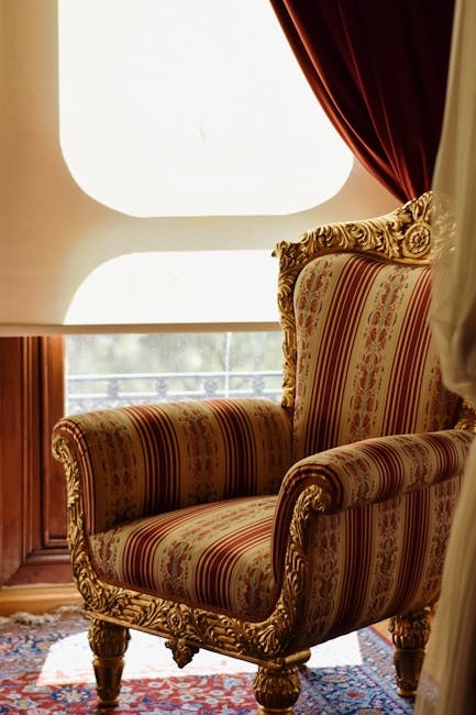 A vintage-style armchair with ornate wooden carvings and striped upholstery featuring red, gold, and cream tones is situated in a well-lit room near a large window. The window has a curved, semi-opaque white shade that diffuses natural light, casting a soft glow across the scene. To the right, a dark red curtain is partially drawn back, revealing a glimpse of the outside. The armchair is placed on a patterned rug with intricate designs, and the surrounding wooden window frame adds warm undertones to the setting. The area appears tidy and clean, reflecting careful domestic cleaning and maintenance by Belsize Park Cleaners as part of their upholstery cleaning and curtain care services for Eton Avenue in NW3, Belsize Park.
