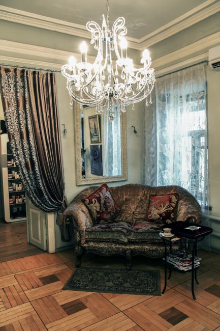 A beige and light green upholstered armchair with gold-colored metal armrests and legs, positioned on a polished wooden herringbone-patterned floor next to a window dressed with sheer white curtains and heavier brown drapes. Natural light filters through the curtains, illuminating the clean and well-maintained domestic room. The setting suggests a focus on home surface cleaning and furniture maintenance, with the armchair appearing freshly cleaned and free of dust or stains. This scene exemplifies professional upholstery cleaning and window treatment care, as provided by Belsize Park Cleaners, seen in the context of comprehensive domestic cleaning services tied to Eton Avenue upholstery cleaning and curtain care NW3, BELSIZE PARK.